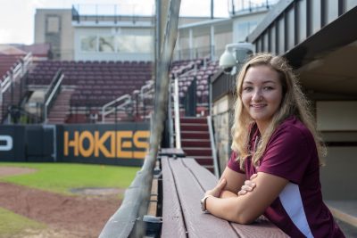 Young woman with blonde hair in a maroon and white Virginia Tech polo on the softball field.