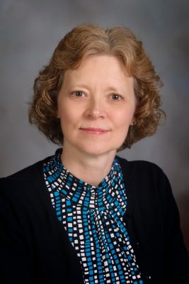 A woman with reddish hair poses in na photo studio, wearing a black blazer over a blue, black, and white checkered shirt.