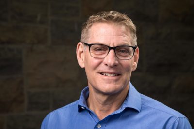 Sporting glasses and bright blue. shirt, a man smiles as he poses for a portrait.