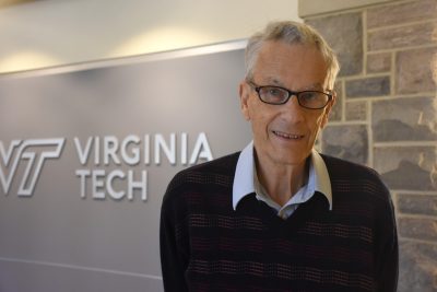 An elderly man with white hair and glasses and wearing a dark sweater stands in front of a wall with Hokie Stone.