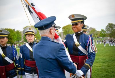  Major General Randal Fullhart hands Cadet Johnson a flag on the Drillfield. Both are in dress uniforms and cadets are in the background. Johnson is smiling.